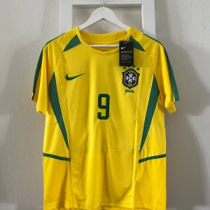 Brasilien Ronaldo 9 fotbollströja Nike - Gul Brasilien fotbollströja från Nike med gröna detaljer, nummer 9 och Ronaldo på ryggen. Broderat CBF-emblem på bröstet och klassisk Nike-logga. Tillverkad i lätt och ventilerande polyester, perfekt för dig som älskar fotboll och vill sticka ut.