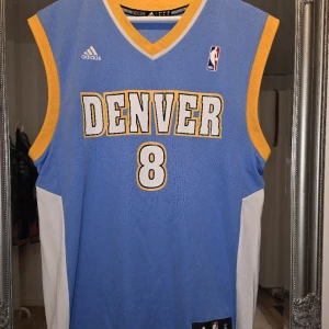 Denver Nuggets Gallinari basketlinne/ NBA JERSEY - Säljer ett blått Denver Nuggets basketlinne från Adidas med Gallinari och nummer 8 på ryggen. Linnet har gula och vita detaljer, ärmlös design och officiella NBA-loggor. Perfekt för basketfans som vill sticka ut på planen eller på läktaren.