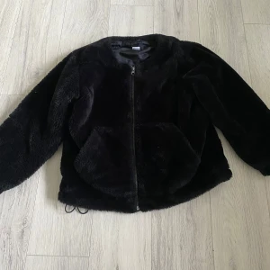 Svart teddyjacka med dragkedja - Mysig svart teddyjacka med dragkedja framtill och stora fickor. Jackan har rund halsringning och långa ärmar, perfekt för kyliga dagar. Den fluffiga pälslooken ger en trendig vibe och passar till många outfits. Dragsko nertill för justerbar passform.