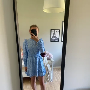 Blå blommig volangklänning - Supersöt blå kortklänning med små vita blommor, volangdetaljer på axlar och nederkant samt långa ärmar med resår vid handlederna. Klänningen har markerad midja och rund halsringning. Perfekt för vår och sommar!