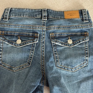 Blå jeans från Perfect Jeans, stl 34 - Snygga blå jeans från Perfect Jeans med klassisk femficksdesign och kontrastsömmar. Byxorna har raka ben, knappar på bakfickorna och en vit snörning i midjan istället för dragkedja. Perfekta för en avslappnad och cool look.