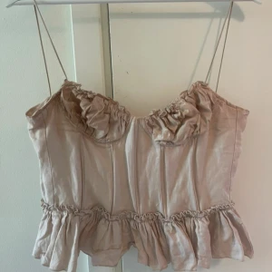 Beige korsettopp med volangdetaljer - Supersöt beige korsettopp med tunna axelband och volangdetaljer både vid bysten och nederkanten. Toppen har en feminin och romantisk vibe med markerade sömmar och lätt rynkade partier. Perfekt att styla med jeans eller kjol för en trendig look. Korsett liknande