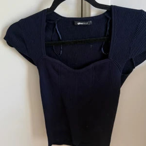 Mörkblå ribbad topp Gina Tricot - Supersnygg mörkblå ribbad topp från Gina Tricot med korta ärmar och söt v-ringning. Toppen har en figurnära passform och är gjord i stretchigt material som sitter perfekt. Perfekt att styla med jeans eller kjol för en trendig look.