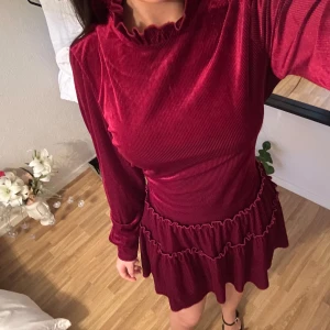 Vinröd sammetsklänning med volang - Supersöt vinröd kort sammetsklänning med långa ärmar, volangdetaljer och hög krage. Klänningen har ett matchande bälte i midjan som knyts och ger en snygg siluett. Perfekt för dig som vill sticka ut med en lyxig och trendig look.