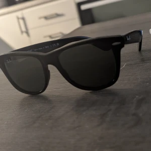 Svarta Ray-Ban Wayfarer solglasögon - Klassiska svarta Wayfarer solglasögon från Ray-Ban. Snygg och tidlös design med svarta bågar och mörka glas. Perfekta för att ge din stil en cool edge och skydda ögonen mot solen.