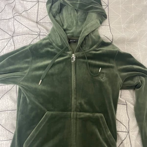 Grön velourjacka från Juicy Couture XS - Mysig grön velourjacka från Juicy Couture med huva, dragkedja och känguruficka. Klassisk JC-logga broderad på bröstet och snörning i huvan. Perfekt för en chill och trendig look.