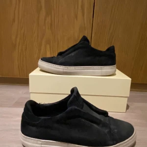 Svarta sneakers från Axel Arigato mocka - Stilrena svarta sneakers från Axel Arigato i mjuk mocka med vit platt sula. Skorna har minimalistisk design utan snörning och en rund tå. Perfekta för dig som gillar en clean och modern look. Dock har sulan hål i hälen men det går att fixa med lim om man vill det👍. Storleken är 43 men passar både dig som har 43 och 44.