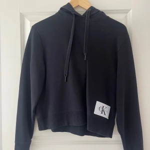 Svart hoodie från Calvin Klein Jeans - Svart hoodie från Calvin Klein Jeans med stor vit logopatch framtill. Tröjan har huva med snörning, ribbade muddar och är croppad i modellen. 