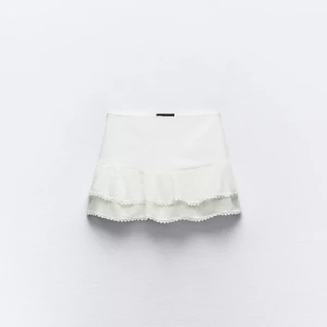 Vit volangkjol med spetsdetaljer - Supersöt vit kortkjol med dubbla volanger och fina spetsdetaljer längst ner. Kjolen har en feminin och fräsch look, perfekt för dig som gillar en ljus och somrig stil. Sitter snyggt i midjan och ger en lekfull vibe.