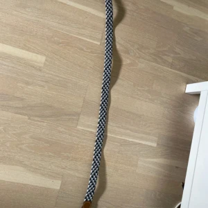 Randigt flätat bälte med skinn-detaljer - Snyggt flätat bälte i svart, vitt och grått med bruna skinn-detaljer och silverspänne. Bältet har ett randigt mönster och är gjort i en mix av textil och skinn, vilket ger en cool och modern look. Perfekt för att lyfta vilken outfit som helst.