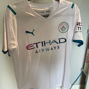 Manchester City Foden 47 bortatröja Puma - Manchester City bortatröja med Foden 47-tryck. Vit med turkosa detaljer och klubbmärke på bröstet. Puma-logga, Etihad Airways sponsortryck och ventilerande funktionsmaterial. Klassisk passform och korta ärmar, perfekt för fotbollsfans.