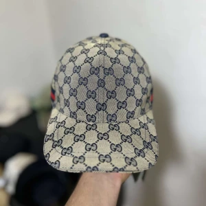 Gucci keps med GG-mönster och röd-blå rand - Snygg keps från Gucci i beige med klassiskt GG-monogram över hela kepsen. Kepsen har en böjd skärm och en ikonisk röd-blå rand på sidan. Tillverkad i bomullsmix och har justerbar rem baktill. Perfekt för dig som vill sticka ut på sommaren☀️