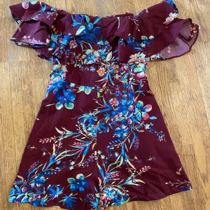 Blommig vinröd kortklänning Luc Ky Star Small - Vinröd byxdress med ett blommigt mönster i blått, rosa och grönt. Den har en volangdetalj runt axlarna och en dragkedja baktill. ﻿Uppskattad storlek S L: ca 68 cm ﻿Bystvidd: 45 cm Polyester.  Inga bilder när den sitter på mig! Katter finns i hemmet!