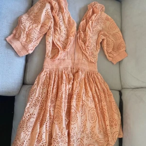 Persikofärgad/korall/orange  kortklänning med volang - Superfin persikofärgad kortklänning i bomull med broderade hålmönster och volangdetaljer längs den djupa v-ringningen. Klänningen har trekvartsärm med breda muddar och markerad midja. Perfekt för sommarens alla tillfällen!