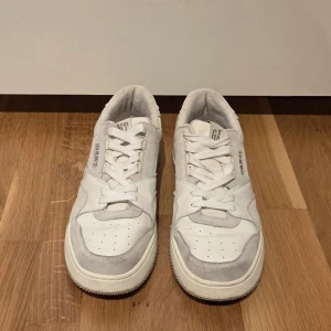 Vita sneakers från Gant i skinn och mocka - Snygga vita sneakers från Gant med detaljer i ljusgrå mocka och skinn. Klassisk låg modell med rund tå, perforerade partier och tydlig logga på sidan och plös. Skorna har snörning och en platt, mönstrad sula för extra grepp.