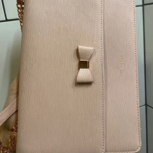 Ljusrosa kuvertväska från Ted Baker - Supersöt ljusrosa kuvertväska från Ted Baker med elegant rosettdetalj framtill och guldfärgad kedja. Väskan är i strukturerat syntetmaterial och har ett mönstrat innerfoder med små rosetter samt praktiskt innerfack med dragkedja. Perfekt för dig som gillar stilrena accessoarer. Äkta läder. 