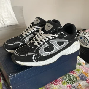 Svarta Dior B30 sneakers med silverdetaljer - Snygga Dior B30 sneakers i svart mesh och skinn med silvriga detaljer och tydlig CD-logga på sidorna och hälen. Skorna har vit och svart chunky sula, vita skosnören och en sportig vibe. Perfekta för dig som vill sticka ut med exklusiva och trendiga sneakers.