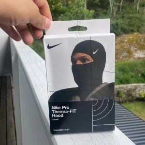 Nike Pro Therma-FIT Hood svart - Nike Pro Therma-FIT Hood är en svart, tight balaclava med minimalistisk design och diskret Nike-logga. Tillverkad i polyester och spandex för stretch och värme, perfekt för fotboll eller träning utomhus. Kan bäras under hjälm eller för sig själv.