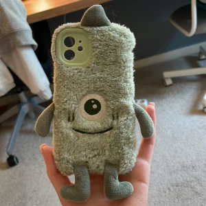 Grön fluffig monster mobilskal - Säljer ett gulligt mobilskal i ljusgrön plysch med ett broderat monsteransikte, ett stort öga och mjuka armar och ben. Skalet är mjukt och har en unik, lekfull design som verkligen sticker ut. Passar perfekt för dig som vill ha något annorlunda till mobilen.