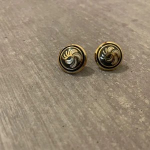 Runda örhängen i svart och guld - Säljer ett par runda örhängen med svart bas och guldfärgad kant. Framsidan har ett snirkligt mönster i silver och guld som ger en cool retrovibe. Perfekta för dig som gillar statement-accessoarer med unik design.