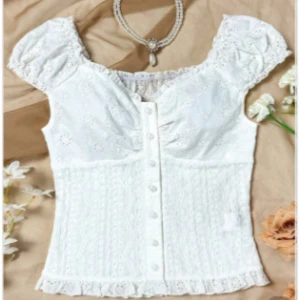 Vit blommig blus med spets och knappar - Supersöt vit blus från Soleia med broderade blommor, spetsdetaljer och volangkant nertill. Blusen har kort puffärm, knappar framtill och v-ringning. Perfekt för dig som gillar romantisk och feminin stil.