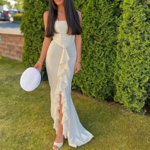 Champagnefärgad långklänning med volang - Elegant långklänning i champagnefärg med figurnära passform och snygg volangdetalj framtill. Klänningen är ärmlös med rak urringning och har en slits framtill. Perfekt för dig som vill sticka ut med en stilren och lyxig look.