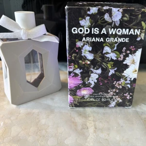 God Is A Woman parfym Ariana Grande - Parfymen God Is A Woman av Ariana Grande, 50 ml. Flaskan är lila med vit kork och dekorerad med en vit rosett. Yttre fodralet är geometriskt och grått, och kartongen har ett blommigt mönster i lila, vitt och grönt. Doften är fräsch och modern.