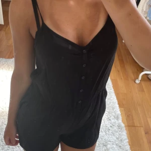 Svart jumpsuit - Svart snygg jumpsuit från Hm med smala axelband och knappar framtill. Jumpsuiten har en avslappnad passform, markerad midja och är tillverkad i mjuk bomull. Perfekt för varma dagar när du vill ha något enkelt men snyggt.