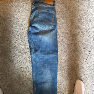 Blå jeans från Jack and Jones - Säljer ett par klassiska blå jeans i rak modell med snyggt slitna detaljer och kontrastsömmar. Jeansen har fem fickor och normal passform. Perfekta till sneakers eller boots för en chill look.