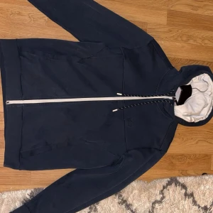Svart hoodiejacka från Hugo Boss - Svart hoodiejacka från Hugo Boss med vit dragkedja och snörning. Jackan har huva med BOSS-logga runt kanten och broderad logga på bröstet. Tillverkad i mjuk bomull, perfekt för en chill och stilren look.