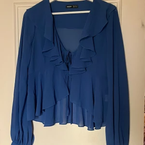 Söt blå blus med volanger - Supersöt blå blus med volanger och knytning framtill. Blusen har även knappar nedtill på armarna som går att knäppa upp. Passar perfekt till en sommarkväll eller som cover up till stranden.