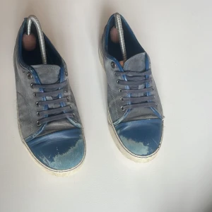 Gråblå sneakers från Lanvin i mocka - Snygga sneakers från Lanvin i grå mocka med blå detaljer och tåparti i blått skinn. Klassisk rund tå, platt sula och snörning. Perfekt för dig som gillar stilrena men ändå unika skor med premiumkänsla.