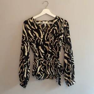 Zebramönstrad omlottblus från Object - Snygg blus från Object i svart och beige zebramönster. Blusen har långa ärmar, knytband i midjan och v-ringning. Materialet är mjukt och har en lätt glansig finish som ger en lyxig känsla. Den har en liten fläck som man ser på bild 4, den smälter dock in i mönstret. Har inte försökt få bort den så mycket möjligt att det går.