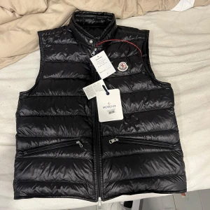 Moncler väst  - Svart dunväst från Moncler med glansig finish och klassisk ståkrage. Västen har två snedställda fickor med dragkedja och Moncler-logga på bröstet. Fylld med vitt gåsdun för extra värme och lätt känsla. Perfekt för lager-på-lager-stil.