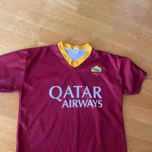 AS Roma Džeko matchtröja röd/gul - AS Roma fotbollströja med Džeko 9 på ryggen. Djup röd färg med gula detaljer och klubbmärke på bröstet. Kortärmad modell i lätt och ventilerande polyester. Snyggt mönster över hela tröjan och Qatar Airways som sponsor framtill.