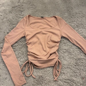 Ribbad rosa långärmad topp med snörning - Supersnygg ribbad topp i ljusrosa med långa ärmar och trendig snörning i sidorna. Toppen har en bred, fyrkantig urringning och croppad passform som ger en modern vibe. Perfekt att styla med jeans eller kjol för en cool look.