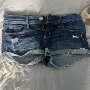Blå jeansshorts från Hollister - Snygga blå jeansshorts från Hollister med låg midja och rullade benslut. Shortsen har slitna detaljer framtill och bakfickor med dekorativa sömmar. Perfekta för sommaren och ger en avslappnad vibe.