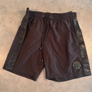 Svarta Balmain badbyxor  - Snygga svarta badshorts från Balmain med breda svarta sidoband där det står 'BALMAIN PARIS' i glansig text. Shortsen har elastisk midja med snörning och en rund Balmain-logga framtill. Perfekta för dig som gillar exklusiv streetstyle. Kvitto finns. 