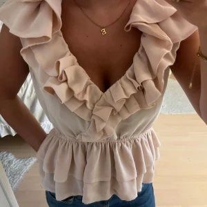 Rosa volangblus med omlott - Supersöt rosa blus med omlott och djup v-ringning. Toppen har flera lager volanger både fram och bak samt markerad midja med resår. Ärmlös modell i ett lätt, flowy material som ger en romantisk vibe. Perfekt att styla med jeans eller kjol för en trendig look.