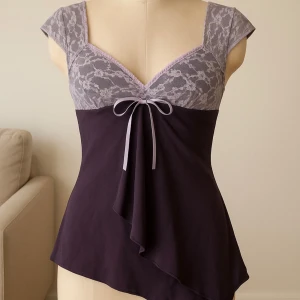 Lila spetslinnetopp med rosett - Supersöt lila linnetopp med ljuslila spets upptill och en liten rosett framtill. Toppen har v-ringning, cap sleeve och asymmetrisk volang nertill. Perfekt för dig som gillar romantiska detaljer och en feminin look.