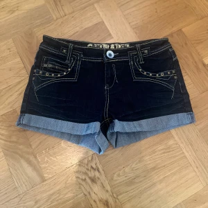 Mörkblå jeansshorts med nitar - Snygga mörkblå jeansshorts från Generation med uppvikta ben, kontrastsömmar och coola nitar på fickorna. Shortsen har klassisk femficksdesign, dragkedja och knapp framtill samt dekorativa detaljer på bakfickorna. Perfekta för sommaren!