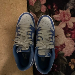 Nike Dunk Low blå och vita sneakers - Nike Dunk Low sneakers i blått och vitt med klassisk Swoosh på sidorna. Skorna har snörning, platt sula och rund tå. Ovandelen är i syntet och skinn, med vit bas och blå paneler. Perfekta för dig som gillar streetwear och vill sticka ut med en ikonisk modell.