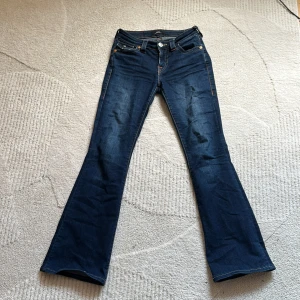 True Religion Becca Bootcut Jeans - Snygga mörkblå jeans från True Religion, modell Becca Mid Rise Bootcut. Klassisk femficksdesign med markerade sömmar och coola fickdetaljer bak. Jeansen har medelhög midja och är tillverkade i stretchig denim för skön passform.