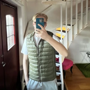 Grön dunväst från Riley - Säljer en snygg grön dunväst från Riley. Västen har en dragkedja framtill och är perfekt för lager-på-lager under kyligare dagar. Den quiltade designen ger en stilren look. Passar bra till både casual och mer uppklädda tillfällen.