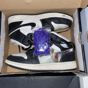 Air Jordan 1s hologram - Snygga svartvita Nike Air Jordans med en unik design på sidan. Skorna har klassisk snörning och kommer med extra lila skosnören. Perfekta för dig som vill ha en stilren men ändå iögonfallande look.