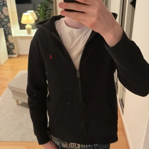 Svart hoodie från Ralph Lauren - Säljer en stilren svart hoodie från Ralph Lauren med dragkedja och det klassiska röda logotypen på bröstet. Perfekt för en avslappnad look. Den har långa ärmar och en bekväm passform.