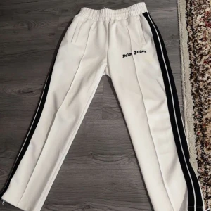  Palm Angels - Snygga vita byxor från Palm Angels med svarta sidoremsor och elastisk midja. Byxorna har en avslappnad passform och är perfekta för en trendig look. Märkesloggan är broderad på framsidan.