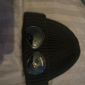 Svart stickad mössa från C.P. Company - Snygg svart stickad mössa från C.P. Company med unika glasögondetaljer på framsidan. Perfekt för att hålla värmen med stil. Märkesetikett på insidan. Nytt skick