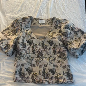 Topp från Neo Noir - Säljer en elegant topp från Neo Noir med v-ringning och korta ärmar. Toppen har ett vackert mönster i guld och svart på en ljus bakgrund. Perfekt för att ge en touch av glamour till din outfit. Aldrig använd så är i jättebra skick. Storlek Men funkar i s också. Pris kan diskuteras 💗