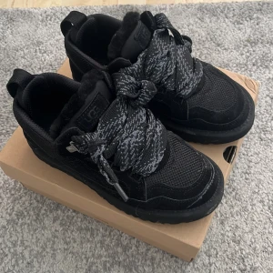 Svarta sneakers från UGG - Snygga svarta sneakers från UGG med tjocka, mönstrade snören och en robust sula. Skorna har en mysig insida och är perfekta för en cool och bekväm stil. De är tillverkade i en kombination av syntet och mocka.